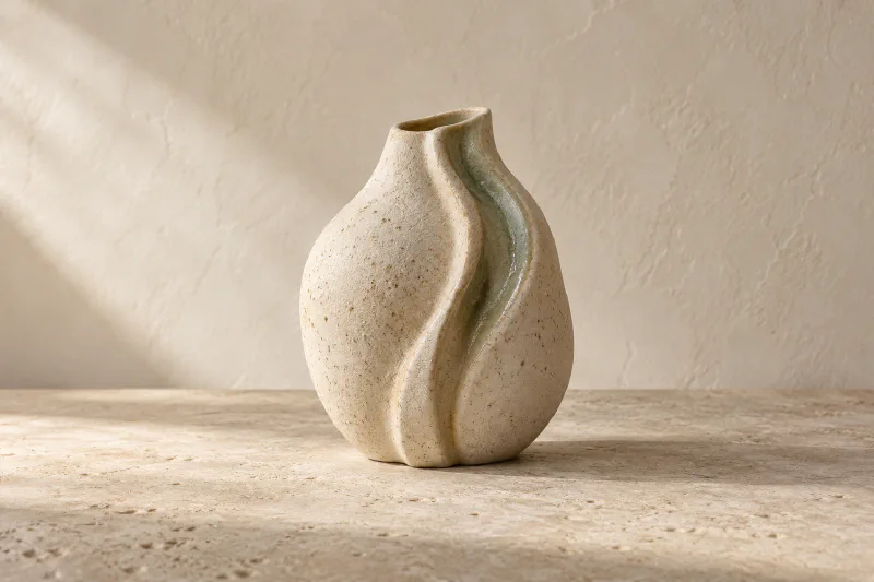 Vase de grès fictif Sillage, ivoire moucheté, relief plissé et glaçure vert sauge sur une surface en pierre.
